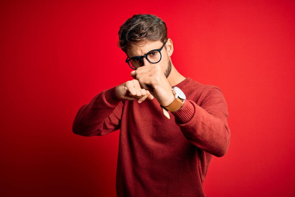 Young handsome man with beard wearing glasses and sweater standing over red background Punching fist to fight, aggressive and angry attack, threat and violence - Photo, image