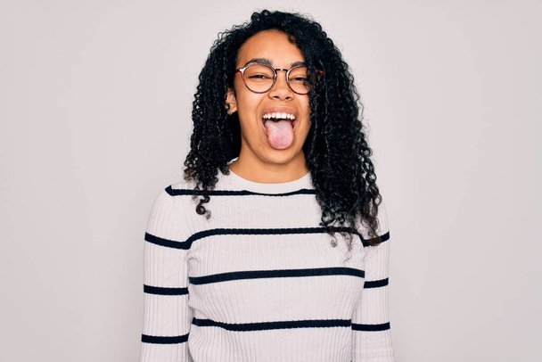 Young african american woman wearing striped sweater and glasses over white background sticking tongue out happy with funny expression. Emotion concept. - Fotografie, Obrázek