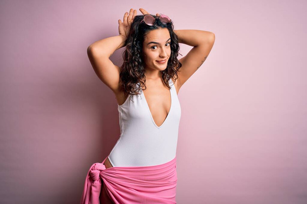 Beautiful woman with curly hair on vacation wearing white swimsuit over pink background Doing bunny ears gesture with hands palms looking cynical and skeptical. Easter rabbit concept. - Фото, изображение