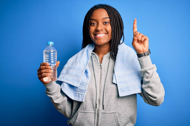 若いアフリカ系アメリカ人のフィットネス女性がタオルを着用し、幸せな顔、ナンバーワンで指を指しているアイデアや質問に驚いてペットボトルから水を飲む - 写真・画像