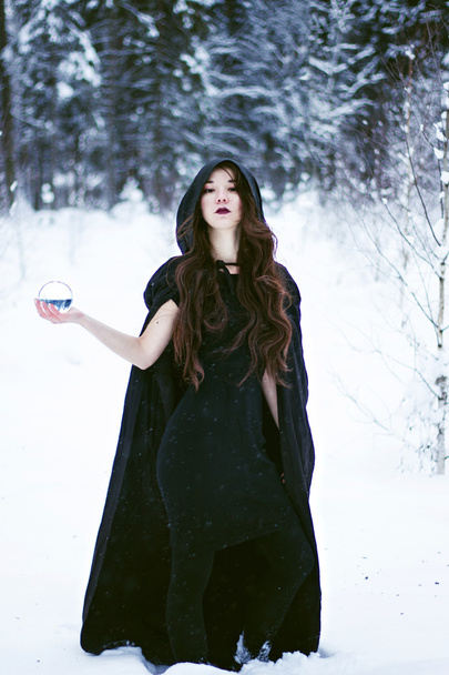 μάγισσα ή γυναίκα σε μαύρη κάπα με γυάλινο σφαιρίδιο στο λευκό χιόνι δάσος - Φωτογραφία, εικόνα