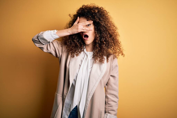 Young beautiful brunette woman with curly hair and piercing wearing casual t-shirt and diadem peeking in shock covering face and eyes with hand, looking through fingers with embarrassed expression. - Фото, изображение