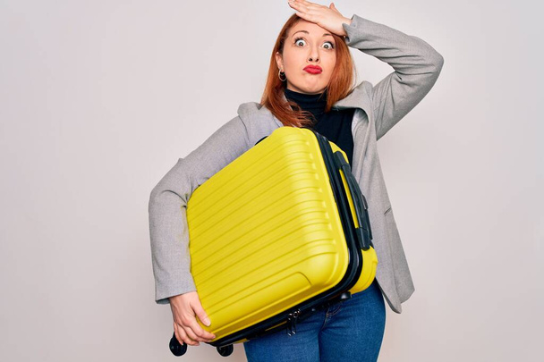 Young beautiful redhead woman holding suitcase prepared to business travel stressed with hand on head, shocked with shame and surprise face, angry and frustrated. Fear and upset for mistake. - Фото, изображение