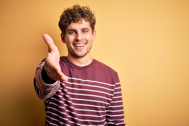 Young blond handsome man with curly hair wearing casual striped sweater smiling friendly offering handshake as greeting and welcoming. Successful business. - Photo, Image