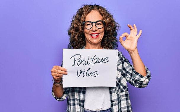 Middle age woman asking for optimistic attitude holding paper with positive vibes message doing ok sign with fingers, smiling friendly gesturing excellent symbol - Photo, Image