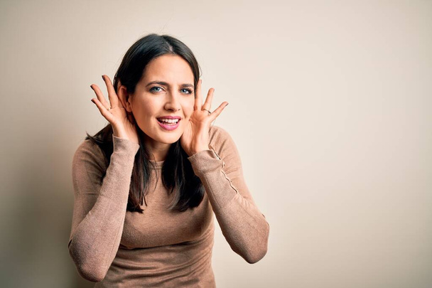 Young brunette woman with blue eyes wearing casual sweater over isolated white background Trying to hear both hands on ear gesture, curious for gossip. Hearing problem, deaf - Photo, Image