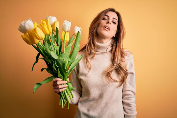 Joven mujer morena hermosa sosteniendo ramo de tulipanes amarillos sobre fondo aislado buscando somnoliento y cansado, agotado por la fatiga y la resaca, ojos perezosos en la mañana
. - Foto, imagen