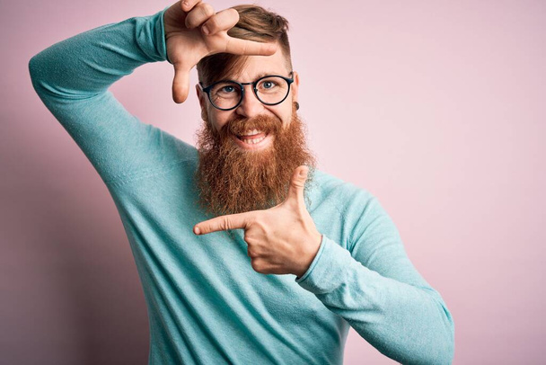 Bella rossa irlandese con barba che indossa occhiali su sfondo rosa isolato sorridente che fa da cornice con le mani e le dita con il viso felice. Concetto di creatività e fotografia. - Foto, immagini