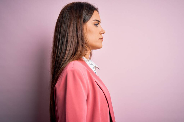 Νεαρή όμορφη μελαχρινή γυναίκα φορώντας σακάκι στέκεται πάνω από απομονωμένο ροζ φόντο κοιτάζοντας προς τα πλάγια, να χαλαρώσετε προφίλ ποζάρουν με φυσικό πρόσωπο με αυτοπεποίθηση χαμόγελο. - Φωτογραφία, εικόνα
