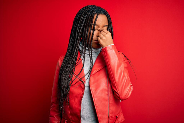 Młoda Afroamerykanka ubrana w skórzaną kurtkę na czerwonym, odizolowanym tle, zmęczona pocieraniem nosa i oczu, zmęczona i z bólem głowy. Koncepcja stresu i frustracji. - Zdjęcie, obraz