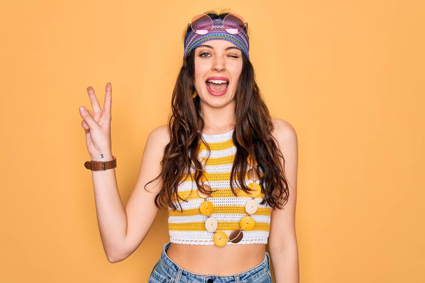 Young beautiful hippie woman with blue eyes wearing accesories and sunnglasses smiling with happy face winking at the camera doing victory sign with fingers. Number two. - Foto, afbeelding