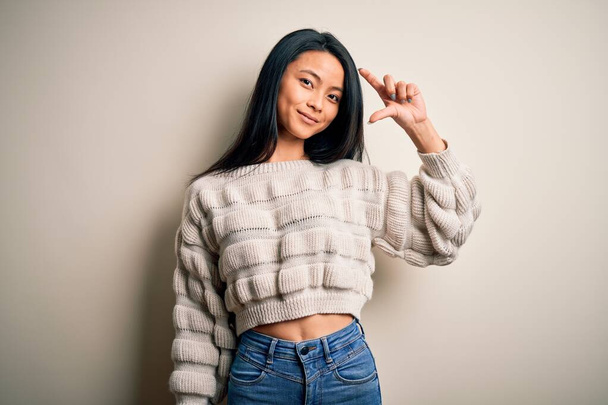 Young beautiful chinese woman wearing casual sweater over isolated white background smiling and confident gesturing with hand doing small size sign with fingers looking and the camera. Measure concept. - Fotó, kép