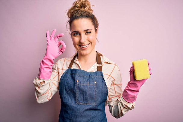 Hermosa mujer rubia limpiadora haciendo tareas domésticas usando arpon y guantes usando scourer haciendo signo de ok con los dedos, símbolo excelente
 - Foto, Imagen