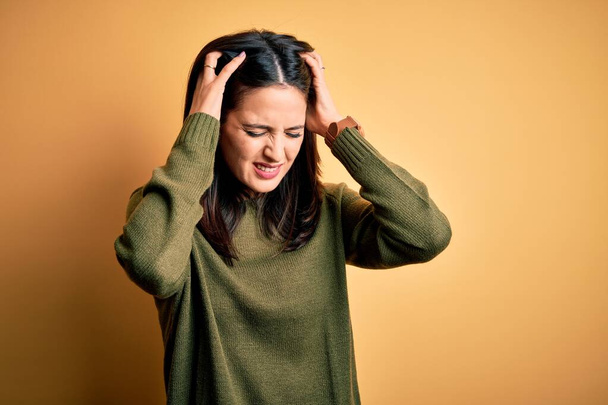 Young brunette woman with blue eyes wearing green casual sweater over yellow background suffering from headache desperate and stressed because pain and migraine. Hands on head. - Foto, afbeelding