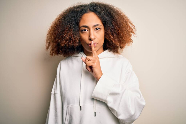 Jonge Afro-Amerikaanse sportvrouw met afrohaar draagt een sportief sweatshirt en vraagt om stil te zijn met de vinger op de lippen. Stilte en geheim concept. - Foto, afbeelding
