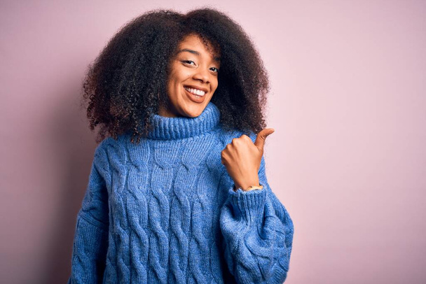 Young beautiful african american woman with afro hair wearing winter sweater over pink background smiling with happy face looking and pointing to the side with thumb up. - Photo, Image