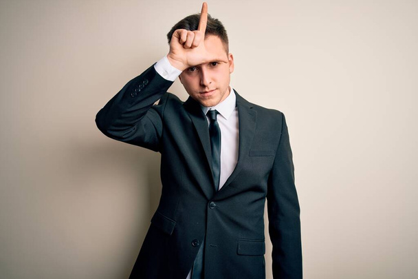 Young handsome business man wearing elegant suit and tie over isolated background making fun of people with fingers on forehead doing loser gesture mocking and insulting. - Foto, Imagen