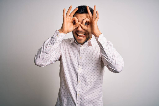Young handsome man wearing elegant shirt standing over isolated white background doing ok gesture like binoculars sticking tongue out, eyes looking through fingers. Crazy expression. - 写真・画像