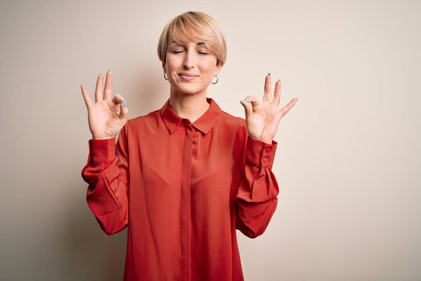 Young beautiful business blonde woman with short hair standing over isolated background relax and smiling with eyes closed doing meditation gesture with fingers. Yoga concept. - Photo, Image