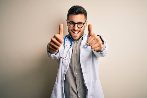 Młody lekarz noszący okulary, medyczny biały szlafrok i stetoskop na odosobnionym tle, aprobujący pozytywny gest ręką, kciuki do góry uśmiechnięte i szczęśliwe dla sukcesu. Gest zwycięzcy. - Zdjęcie, obraz