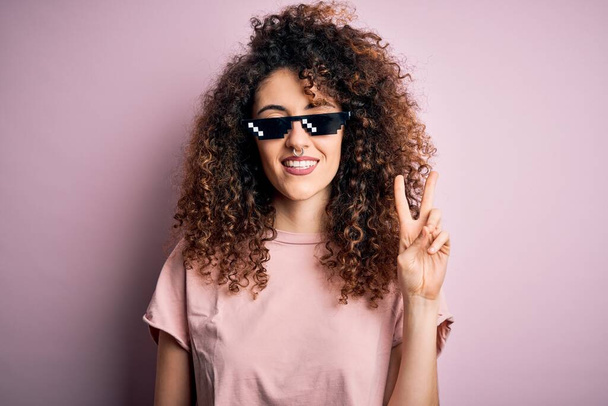 Young beautiful woman with curly hair and piercing wearing funny thug life sunglasses smiling with happy face winking at the camera doing victory sign. Number two. - Foto, imagen