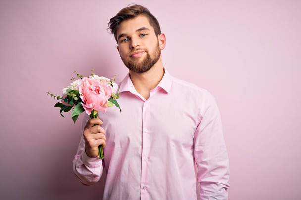 Młody blondyn romantyczny mężczyzna z brodą i niebieskimi oczami trzyma piękny bukiet kwiatów z pewnym siebie wyrazem mądrej twarzy myśląc poważnie - Zdjęcie, obraz