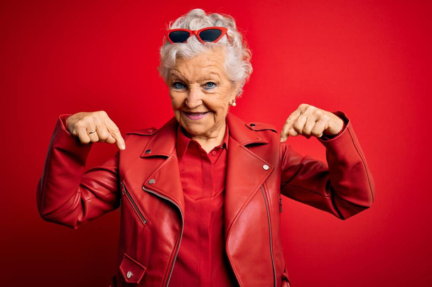 Senior belle femme aux cheveux gris portant une veste rouge décontractée et des lunettes de soleil à l'air confiant avec le sourire sur le visage, se montrant du doigt fier et heureux
. - Photo, image