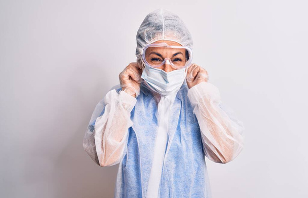 Женщина средних лет носит защитное коронавирусное оборудование на белом фоне, закрывая уши пальцами с раздраженным выражением для шума громкой музыки. Глухая концепция
. - Фото, изображение