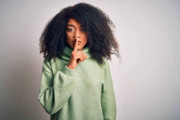 Νεαρή όμορφη Αφροαμερικανή γυναίκα με αφρο μαλλιά που φοράει πράσινο χειμωνιάτικο πουλόβερ ζητώντας να είναι ήσυχη με το δάχτυλο στα χείλη. Σιωπή και μυστική έννοια. - Φωτογραφία, εικόνα