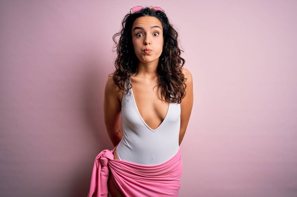 Beautiful woman with curly hair on vacation wearing white swimsuit over pink background puffing cheeks with funny face. Mouth inflated with air, crazy expression. - Zdjęcie, obraz
