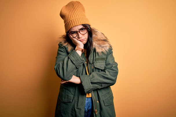 Jeune femme brune portant des lunettes et un manteau d'hiver avec chapeau sur fond isolé jaune pensant fatigué et ennuyé avec des problèmes de dépression avec les bras croisés
. - Photo, image