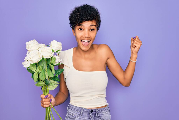 Beautiful african american afro woman holding bouquet of white flowers over purple background screaming proud and celebrating victory and success very excited, cheering emotion - Photo, Image