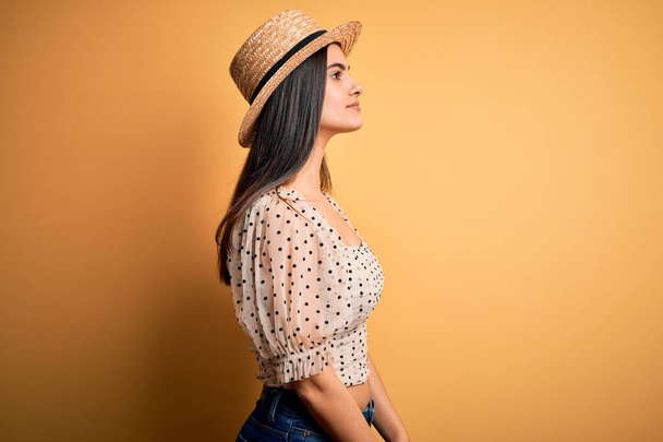 Mladá krásná brunetka žena v tričku a letním klobouku přes žluté pozadí dívá na stranu, relaxační profil póza s přirozenou tváří s sebevědomým úsměvem. - Fotografie, Obrázek