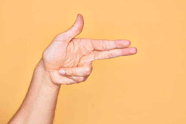 Ręka białego młodzieńca pokazuje palce nad odizolowanym żółtym tłem, gestykulując bronią palną palcami, celując w symbol strzelaniny. - Zdjęcie, obraz