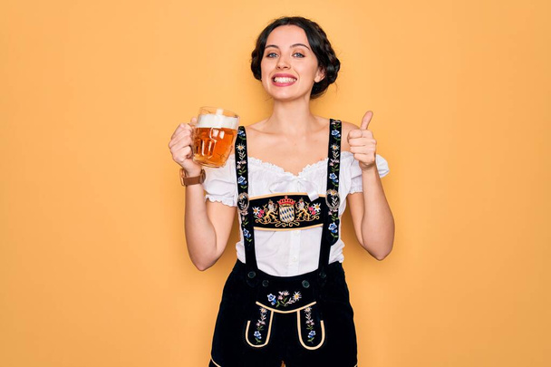 Beautiful german woman with blue eyes wearing traditional octoberfest dress drinking jar of beer happy with big smile doing ok sign, thumb up with fingers, excellent sign - Fotografie, Obrázek