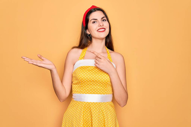 Young beautiful pin up woman wearing 50s fashion vintage dress over yellow background amazed and smiling to the camera while presenting with hand and pointing with finger. - 写真・画像
