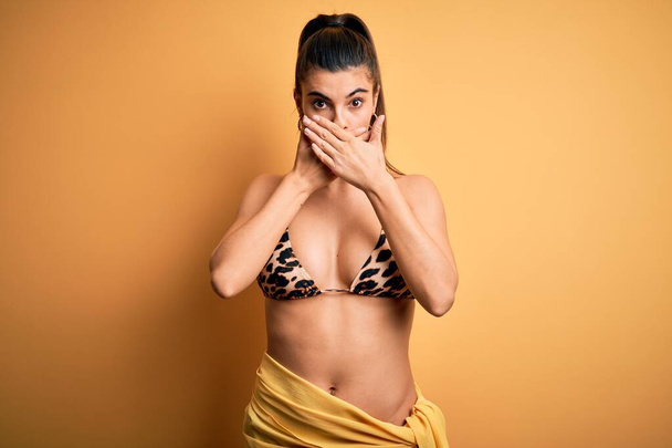 Giovane bella donna bruna in vacanza indossando bikini costume da bagno su sfondo giallo scioccato che copre la bocca con le mani per errore. Concetto segreto
. - Foto, immagini