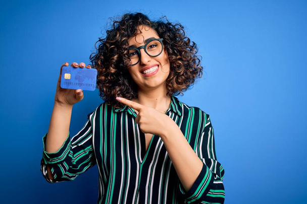 Giovane bella donna araba riccia con gli occhiali che tengono i soldi della carta di credito per il pagamento molto felice puntando con mano e dito
 - Foto, immagini
