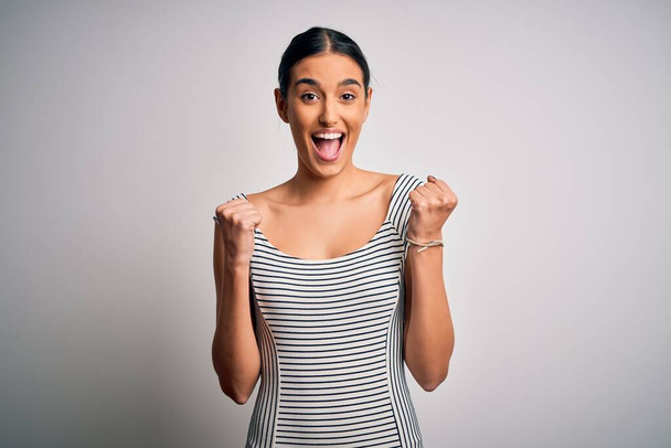 Young beautiful brunette woman wearing casual striped dress over isolated white background celebrating surprised and amazed for success with arms raised and open eyes. Winner concept. - Photo, Image