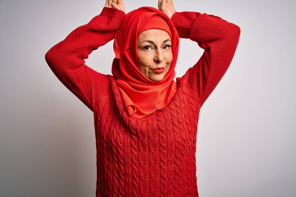Mulher morena de meia idade vestindo muçulmano tradicional hijab sobre fundo branco isolado Fazendo orelhas de coelho gesto com as mãos palmas olhar cínico e cético. Conceito de coelho de Páscoa
. - Foto, Imagem