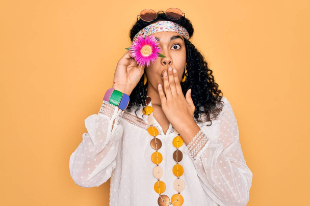 Νεαρή Αφροαμερικανή σγουρή χίπισσα φορώντας γυαλιά ηλίου κρατώντας ροζ λουλούδι στο μάτι στόμα με το χέρι σοκαρισμένη από ντροπή για λάθος, έκφραση φόβου, φοβισμένη στη σιωπή, μυστική έννοια - Φωτογραφία, εικόνα