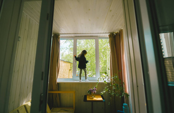 Dívka v lehkých šatech stojí opodál u velkého okna. Koncept karantény během koronaviru. Vodorovná fotografie - Fotografie, Obrázek