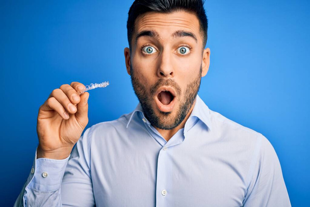 Fiatal jóképű férfi fogja fogászati aligner fogkorrekció felett kék háttér megijedt a sokk egy meglepetés arc, fél, és izgatott a félelem kifejezés - Fotó, kép