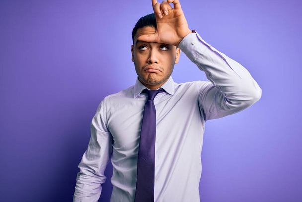Jeune homme d'affaires brésilien portant une cravate élégante debout sur fond violet isolé se moquant des gens avec les doigts sur le front faisant geste perdant moqueur et insultant
. - Photo, image