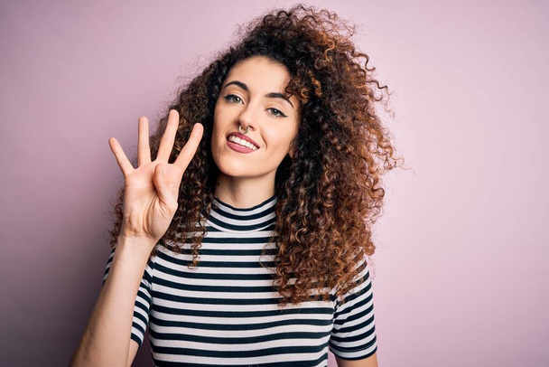 Young beautiful woman with curly hair and piercing wearing casual striped t-shirt showing and pointing up with fingers number four while smiling confident and happy. - Фото, изображение