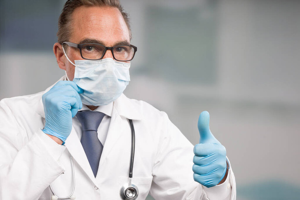 Male family doctor in doctor's overall and tie with stethoscope, medical gloves and medical face mask shows thumbs up - Photo, Image