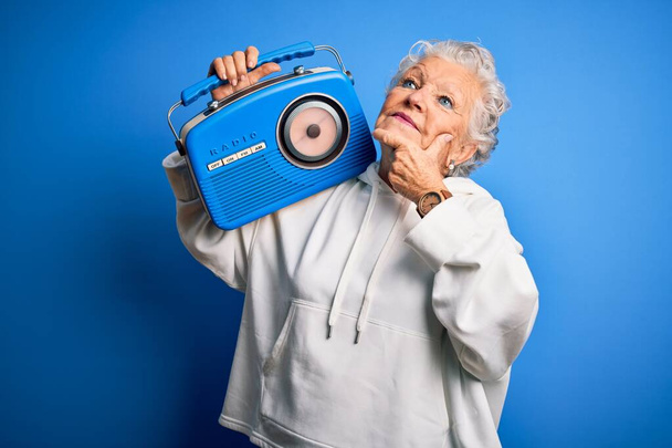 Ältere schöne Frau hält Vintage-Radio über isolierten blauen Hintergrund ernstes Gesicht über Frage nachdenken, sehr verwirrte Idee - Foto, Bild