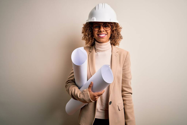Africano americano rizado arquitecto mujer con casco de seguridad y gafas sosteniendo planos con una cara feliz de pie y sonriendo con una sonrisa confiada mostrando los dientes
 - Foto, Imagen