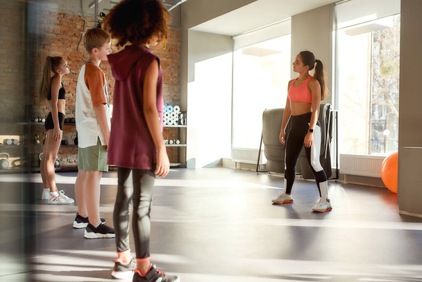 Commit to training. Full-length shot of three teens, boy and girls warming up, exercising with female trainer before workout in gym. Sport, healthy lifestyle, active childhood concept - Photo, Image