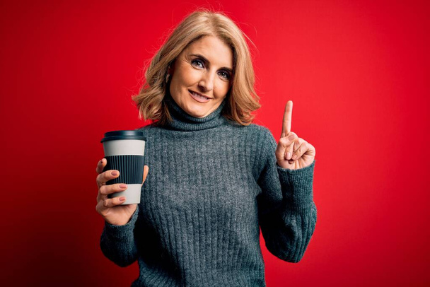 Középkorú gyönyörű szőke nő iszik elvitelre csésze kávét piros háttér meglepett egy ötlet vagy kérdés ujjal mutogató boldog arc, első számú - Fotó, kép